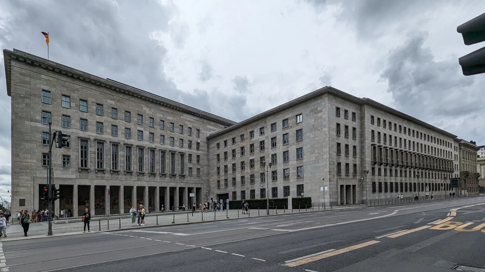 Detlev-Rohwedder-Haus in Berlin, Sitz des Bundesfinanzministeriums: zwei Flügel des langgestreckten Steinbaus an einer Straßenecke unter bewölktem Himmel, davor einzelne Passanten
