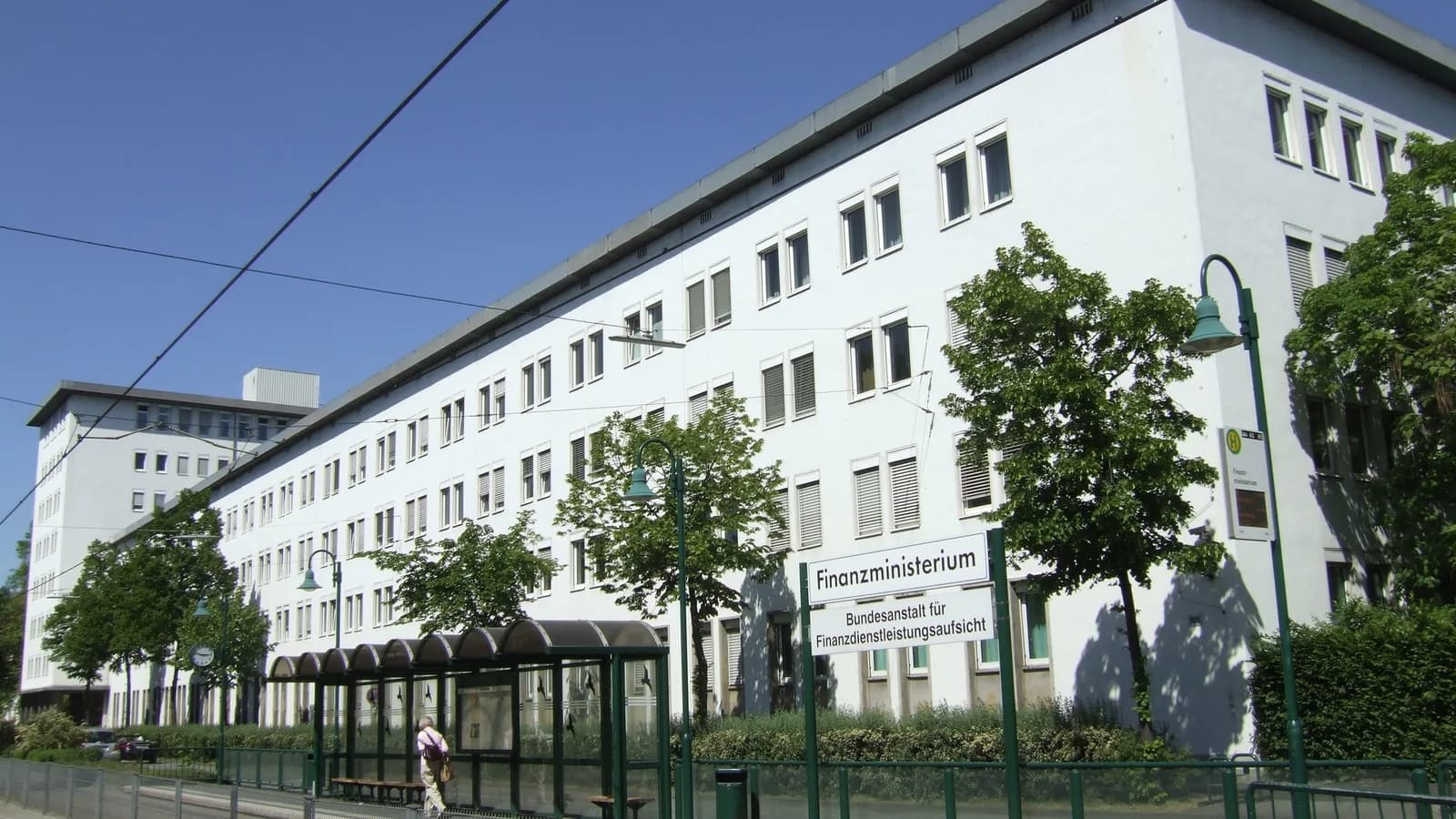 Lange weiße Gebäudefront der Bundesanstalt für Finanzdienstleistungsaufsicht in Bonn unter blauem Himmel, davor Straßenbahnhaltestelle mit Schild, Bäume und eine wartende Person
